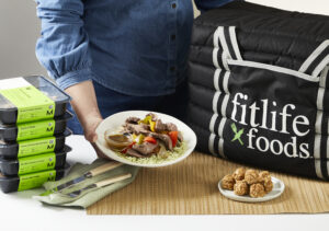 Between a Fitlife Foods cooler bag and a stack of prepared FitMeals, a person holds out Fitlife Foods' Steak Fajita Bowl on a plate